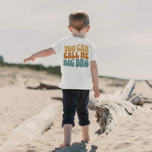 A boy walking on the beach with his back to the camera. On the back of his shirt it reads in big bubbly letters "You can call me big bro". The text is color blocked line by line with the top line in yellow, the second in orange, and the third in teal. 